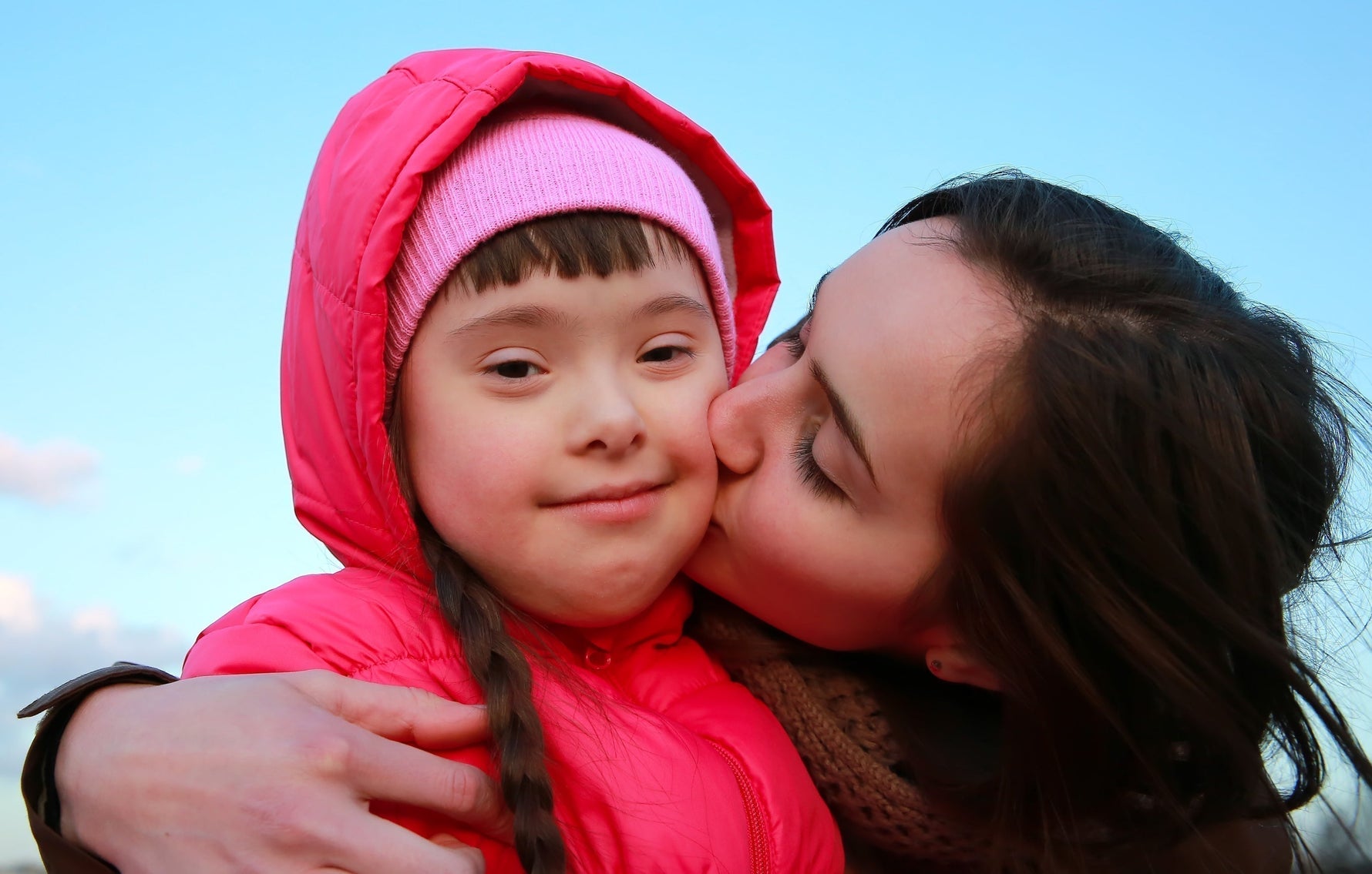 Mother kissing child with Down syndrome on the cheek outdoors in pink winter coats
