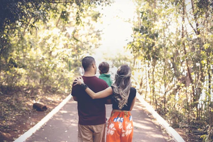 Family of three walking hand-in-hand down a sunny tree-lined path from behind